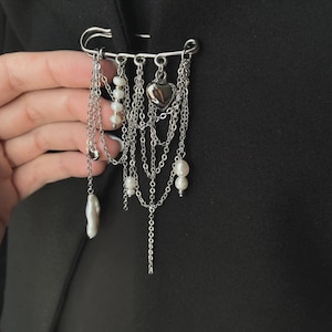 Peut inclure: Une broche épingle à nourrice argentée ornée de multiples chaînes pendantes, de petites perles blanches et d'un cœur en métal argenté poli. La broche est fixée sur un vêtement en tissu noir, présentant un design superposé aux longueurs variées.
