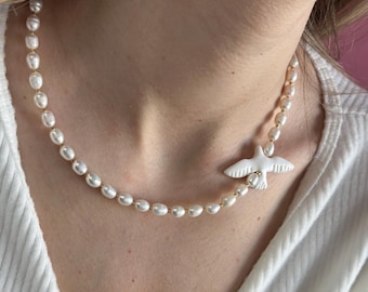 Puede incluir: Un collar que presenta una hilera de pequeñas perlas de agua dulce blancas e irregulares intercaladas con finos espaciadores dorados. Un único dije blanco mate con forma de ave está colocado de forma asimétrica en el lado derecho.