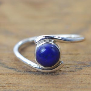 Op de afbeelding: Een zilveren ring met een blauwe lapis lazuli edelsteen. De ring heeft een eenvoudig, modern ontwerp met een gebogen band.
