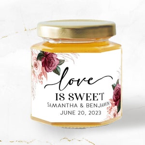 May include: A small glass jar filled with honey, with a white label that reads "love is sweet" and "Samantha & Benjamin June 20, 2023". The label is decorated with pink and red floral illustrations.