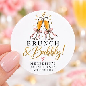 May include: A round white sticker featuring a watercolor illustration of two champagne flutes with pink bows, a small heart, and the text BRUNCH & Bubbly! MEREDITH'S BRIDAL SHOWER APRIL 27, 2025 in black and gold lettering on a clean white background.