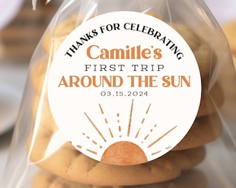 May include: A white circle sticker with the text "Thanks for celebrating Camille's First Trip Around the Sun 03.15.2024" and a brown sun with orange rays. The sticker is on a stack of cookies in a clear plastic bag.