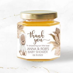 May include: A small glass jar with a white label that says "thank you" for showering our little bear Janna & Rob's Baby Shower 06.15.2023. The jar is filled with honey and has a brown teddy bear with a white hooded sweater on the label.
