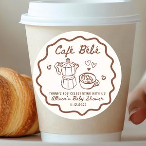 May include: A paper coffee cup with a white lid, featuring a circular sticker that reads "Café Bébé" with a coffee pot and cup illustration. A golden-brown croissant rests on a plate beside the cup. The sticker also includes the text "Thanks for celebrating with us Allison's Baby Shower 11.13.2024".