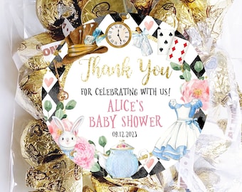 May include: A white and gold thank you favor bag with a round sticker featuring Alice in Wonderland characters and the text "Thank you for celebrating with us! Alice's Baby Shower 08.12.2023".