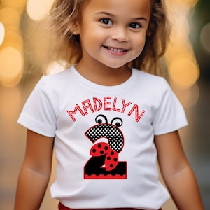 May include: A white t-shirt with a black number two that has red ladybug wings and black polka dots. The shirt also has the name "Madelyn" written in black above the number. The girl is wearing a red bow in her hair and a red skirt.