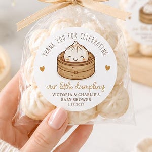 May include: Clear cellophane bags filled with white dumplings, tied with gold ribbon. Each bag has a circular sticker with a dumpling illustration and the text "Thank you for celebrating our little dumpling Victoria & Charlie's Baby Shower 6.14.2027."