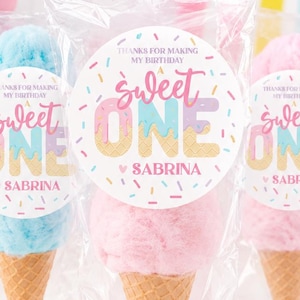 May include: Cotton candy cones in clear cellophane bags. The cotton candy is blue and pink, atop a waffle cone. Each cone has a circular sticker that reads "Sweet One Sabrina" with colorful sprinkles. A bowl of sprinkles and a pink cake stand are in the background.