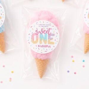 May include: Cotton candy cones in clear plastic bags. The cotton candy is pink and blue, with a cone-shaped waffle cone base. Each bag has a circular sticker that says "Sweet One" and "Sabrina". Sprinkles and straws are also visible.