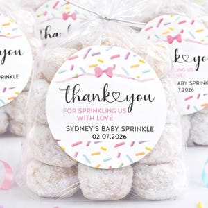 Pode incluir: Várias sacolas transparentes cheias de guloseimas redondas e claras. Cada sacola tem um adesivo circular com o texto "thank you" e "Sydney's Baby Sprinkle 02.07.2026". Balões pastel e confetes no fundo.