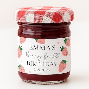 May include: A small glass jar filled with red jam, featuring a red and white checkered lid. The jar has a label with the text "EMMA'S berry first BIRTHDAY 2.15.2026" and strawberry illustrations.
