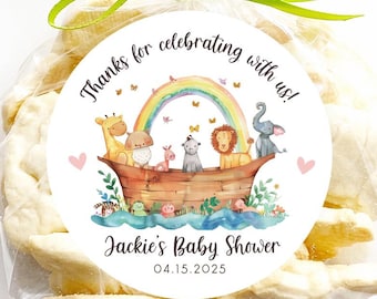 May include: A white favor bag filled with cookies and a round sticker that says "Thanks for celebrating with us! Jackie's Baby Shower 04.15.2023". The sticker features a watercolor illustration of a rainbow, a boat, and animals.