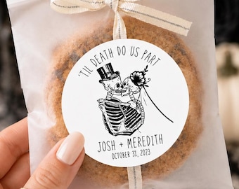 May include: A round cookie inside a translucent white paper bag tied with a silver-trimmed ribbon. A circular white sticker features a black line drawing of two skeletons kissing with the text 'TIL DEATH DO US PART', 'JOSH + MEREDITH', and 'OCTOBER 31, 2023'.