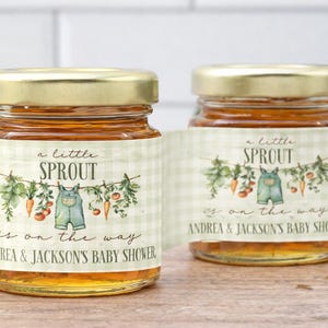 May include: Two glass jars filled with amber liquid, each topped with a gold metal lid. The labels feature a light green gingham pattern, a clothesline illustration with vegetables and baby overalls, and the text A little SPROUT is on the way.