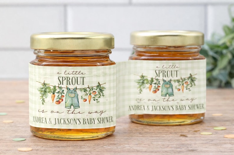 May include: Two glass jars filled with amber liquid, each topped with a gold metal lid. The labels feature a light green gingham pattern, a clothesline illustration with vegetables and baby overalls, and the text A little SPROUT is on the way.