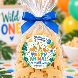 May include: A clear plastic bag filled with animal-shaped crackers, tied with a blue satin ribbon. A circular sticker on the front features a colorful jungle theme with an elephant, giraffe, and lion, text reading "THANKS for coming PARTY ANIMAL! Matthew 05.16.2027".