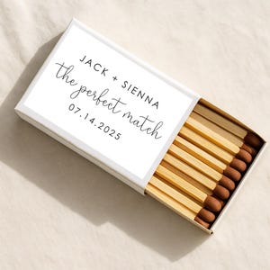 May include: A white matchbox with the text "JACK + SIENNA the perfect match 07.14.2025" in black script. The matchbox is open, revealing wooden matches with brown tips.