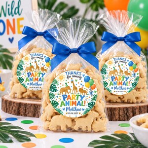 May include: Three clear plastic favor bags filled with animal-shaped crackers, each tied with a blue satin ribbon. A round sticker on each bag features illustrations of a lion, elephant, and giraffe with the text "THANKS for coming PARTY ANIMAL! Matthew 05.16.2027".