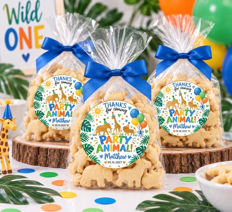 May include: Three clear plastic favor bags filled with animal-shaped crackers, each tied with a blue satin ribbon. A round sticker on each bag features illustrations of a lion, elephant, and giraffe with the text "THANKS for coming PARTY ANIMAL! Matthew 05.16.2027".