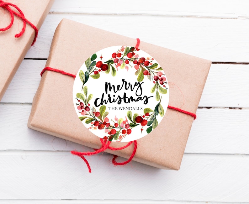 May include: Round white gift tag with a watercolor wreath of red berries and green leaves. The text "Merry Christmas" is written in black cursive lettering with the name "The Wendalls" below.