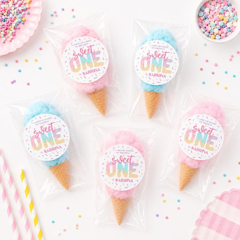 May include: Cotton candy cones in clear plastic bags. The cotton candy is pink and blue, with a cone-shaped waffle cone base. Each bag has a circular sticker that says "Sweet One" and "Sabrina". Sprinkles and straws are also visible.