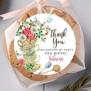 May include: Round white sticker with a watercolor illustration of a stack of teacups with flowers and butterflies. The text on the sticker reads "Thank You for making my party tea-riffic Sabrina".