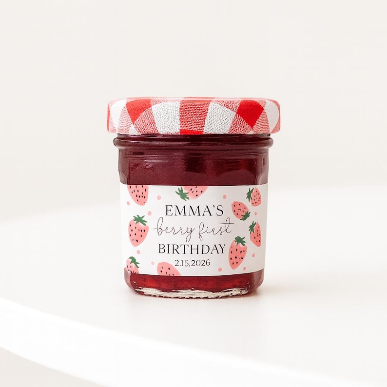 May include: A small jar of jam with a red and white checkered lid. The label reads "EMMA'S berry first BIRTHDAY 2.15.2026" with strawberry illustrations. The jam is a deep red color.