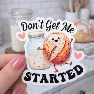 May include: A die-cut sticker featuring a cartoon sourdough starter jar and a smiling, anthropomorphic sourdough bread loaf. The text reads Don't Get Me STARTED in black lettering, accented by two small pink hearts on a white background.