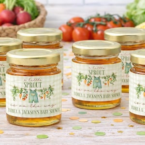 May include: Five small glass jars filled with golden honey, each featuring a label with a green gingham pattern, a watercolor illustration of vegetables and overalls, and the text "a little SPROUT is on the way ANDREA & JACKSON'S BABY SHOWER."