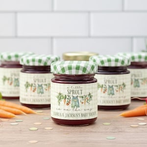 May include: Five glass jars filled with dark red jam, each topped with a green and white checkered lid. The labels feature a garden illustration with carrots, radishes, and overalls, with the text A little SPROUT is on the way.