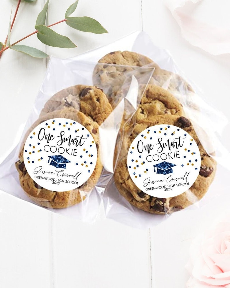 May include: Two clear plastic bags filled with chocolate chip cookies. Each cookie has a round sticker with gold and blue glitter, the text "One Smart Cookie", a graduation cap, and the name "Jessica Criswell" with the text "Greenwood High School 2023".