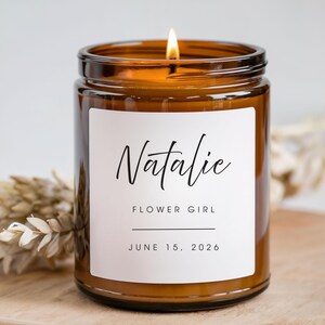 K&ouml;nnte beinhalten: Eine brennende Kerze in einem braunen Glas mit einem wei&szlig;en Etikett. Auf dem Etikett steht "Natalie, Flower Girl, 15. Juni 2026" in schwarzer Schrift. Die Kerze steht auf einer Holzoberfl&auml;che.