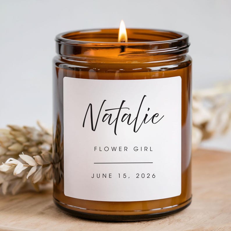 K&ouml;nnte beinhalten: Eine brennende Kerze in einem braunen Glas mit einem wei&szlig;en Etikett. Auf dem Etikett steht "Natalie, Flower Girl, 15. Juni 2026" in schwarzer Schrift. Die Kerze steht auf einer Holzoberfl&auml;che.