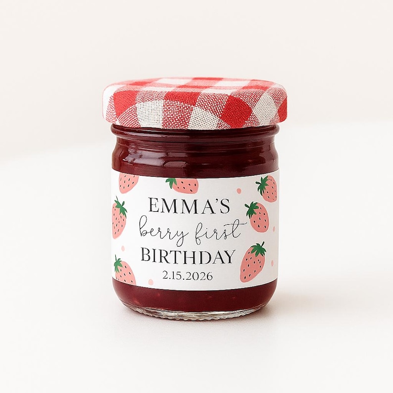 May include: A small glass jar filled with red jam, featuring a red and white checkered lid. The jar has a label with the text "EMMA'S berry first BIRTHDAY 2.15.2026" and strawberry illustrations.