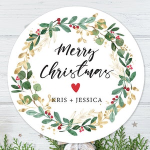 May include: White round Christmas ornament with a green and gold wreath, red berries, and the text "Merry Christmas" with a red heart and "Kris + Jessica" below.