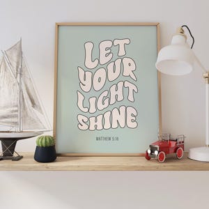 Pode incluir: Um cartaz azul claro com o texto "Let your light shine" em rosa, com um estilo de fonte retr&ocirc;. O texto "Matthew 5:16" &eacute; impresso abaixo do texto principal.