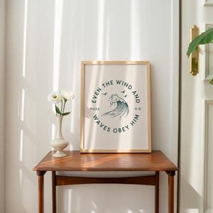 May include: A framed print with a light beige background and a circular design featuring a wave and text that reads "Even the wind and waves obey him." The print is in a wooden frame and sits on a wooden table. A vase with white flowers is next to the frame.