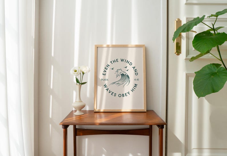 May include: A framed print with a light beige background and a circular design featuring a wave and text that reads "Even the wind and waves obey him." The print is in a wooden frame and sits on a wooden table. A vase with white flowers is next to the frame.