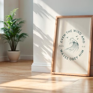 May include: A framed print with a light wood frame, leaning against a white wall. The print features a circular design with the text "EVEN THE WIND AND WAVES OBEY HIM" and a wave illustration. A potted plant is in the background.