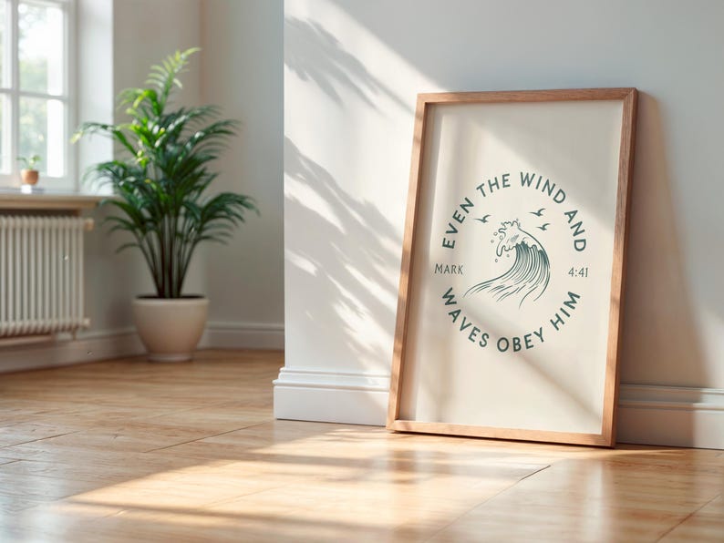 May include: A framed print with a light wood frame, leaning against a white wall. The print features a circular design with the text "EVEN THE WIND AND WAVES OBEY HIM" and a wave illustration. A potted plant is in the background.