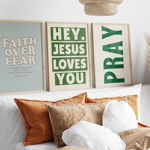 Op de afbeelding: Drie ingelijste kunstprints met inspirerende citaten. De eerste print zegt "Faith Over Fear" in een retro lettertype. De tweede print zegt "Hey Jesus Loves You". De derde print zegt "Pray". De prints worden weergegeven boven een bed met kussens.