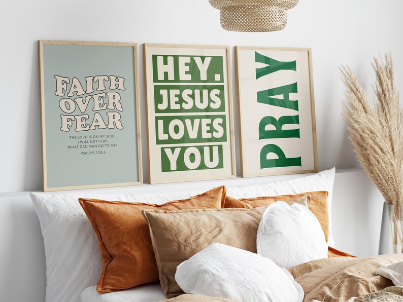 Op de afbeelding: Drie ingelijste kunstprints met inspirerende citaten. De eerste print zegt "Faith Over Fear" in een retro lettertype. De tweede print zegt "Hey Jesus Loves You". De derde print zegt "Pray". De prints worden weergegeven boven een bed met kussens.