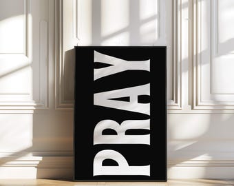 May include: A framed black and white art print. The word "PRAY" is vertically displayed in large, bold, white letters against a black background. The artwork is displayed on a wooden floor, leaning against a white wall.