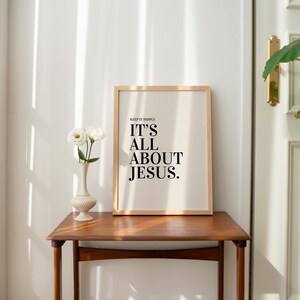 K&ouml;nnte beinhalten: Ein gerahmter Druck mit dem Text "IT'S ALL ABOUT JESUS" in einem Holzrahmen, der auf einem Holztisch steht. Links steht eine Vase mit wei&szlig;en Blumen. Der Raum hat eine neutrale Farbpalette.