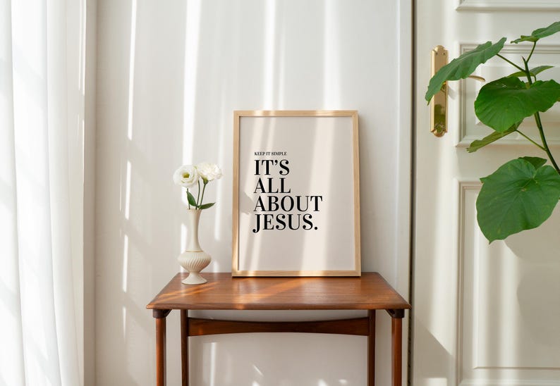 K&ouml;nnte beinhalten: Ein gerahmter Druck mit dem Text "IT'S ALL ABOUT JESUS" in einem Holzrahmen, der auf einem Holztisch steht. Links steht eine Vase mit wei&szlig;en Blumen. Der Raum hat eine neutrale Farbpalette.
