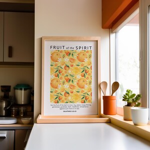 May include: A framed print with a yellow background and a pattern of oranges and white flowers. The print has the text "FRUIT of the SPIRIT" at the top and a Bible verse from Galatians 5:22-23 at the bottom.