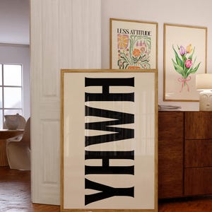 Pode incluir: Uma impressão artística emoldurada com a palavra "YHWY" em letras pretas em negrito sobre um fundo creme. Duas outras impressões artísticas emolduradas são visíveis, uma com o texto "LESS ATTITUDE" e outra com um design floral em aquarela. As impressões são exibidas em uma sala com um armário de madeira.