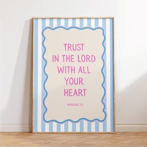 Pode incluir: Uma impressão emoldurada com o texto "TRUST IN THE LORD WITH ALL YOUR HEART" em rosa, sobre um fundo bege com uma borda azul ondulada. A moldura tem um padrão de listras azuis e brancas.