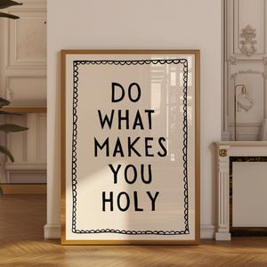 Puede incluir: Una impresión enmarcada con el texto "DO WHAT MAKES YOU HOLY" en letras negras en negrita sobre un fondo crema. El marco es de color marrón claro y la impresión tiene un borde negro decorativo.