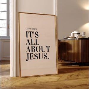 K&ouml;nnte beinhalten: Ein gerahmter Druck mit dem Text "KEEP IT SIMPLE IT'S ALL ABOUT JESUS." Der Druck hat einen goldenen Rahmen und lehnt an einem wei&szlig;en T&uuml;rrahmen. Der Hintergrund umfasst einen Holzboden und einen Holzschrank.